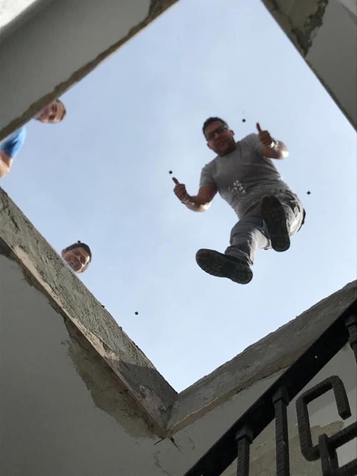 Persona saltando desde un borde en construcción hacia la cámara, con dos hombres observando desde el mismo borde. Cielo despejado de fondo.