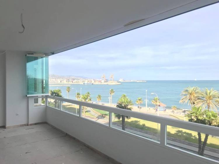 Balcón con piso sin terminar, ventanas de cristal corredizo y una vista al mar con palmeras, un paseo marítimo y un puerto en el horizonte.